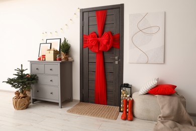 Wooden door with beautiful red bow near chest of drawers, evergreen trees and gift boxes indoors. Christmas decoration