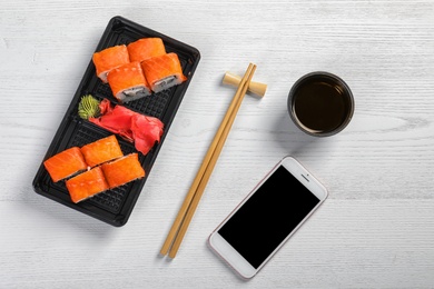 Photo of Flat lay composition with sushi rolls, smartphone and space for text on white wooden table. Food delivery