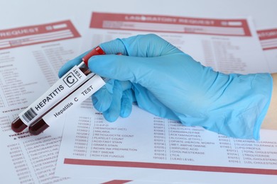 Scientist holding tubes with blood samples for hepatitis virus test against laboratory forms, closeup