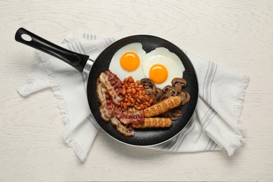 Photo of Frying pan with cooked traditional English breakfast and napkin on white wooden table, top view