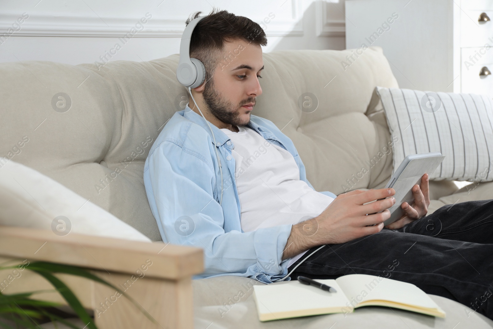 Photo of Young man with headphones using modern tablet for studying on sofa at home. Distance learning