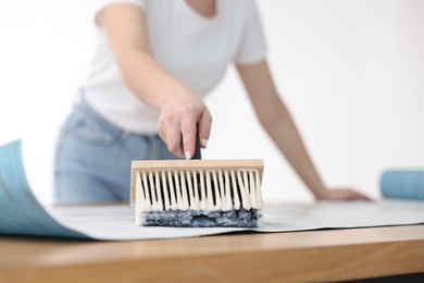 Woman applying glue onto wallpaper sheet at table indoors, closeup