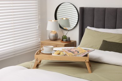 Photo of Wooden tray table with cup of drink and magazines on bed near window with horizontal blinds in room