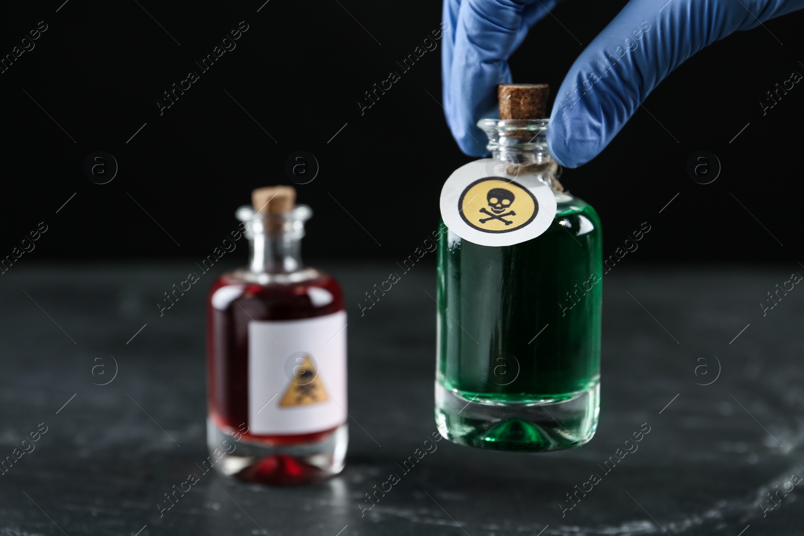 Photo of Woman in gloves holding glass bottle of poison with warning sign over table, closeup. Space for text