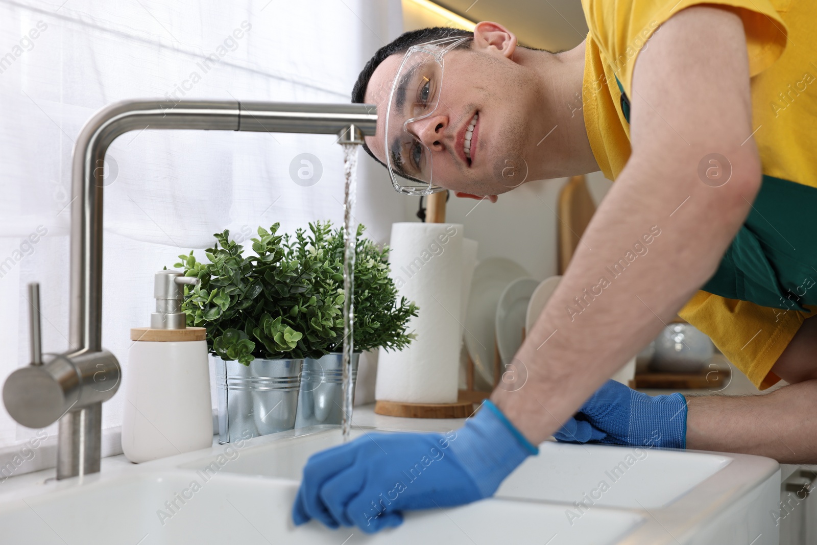 Photo of Smiling plumber wearing protective glasses and gloves examining faucet in kitchen