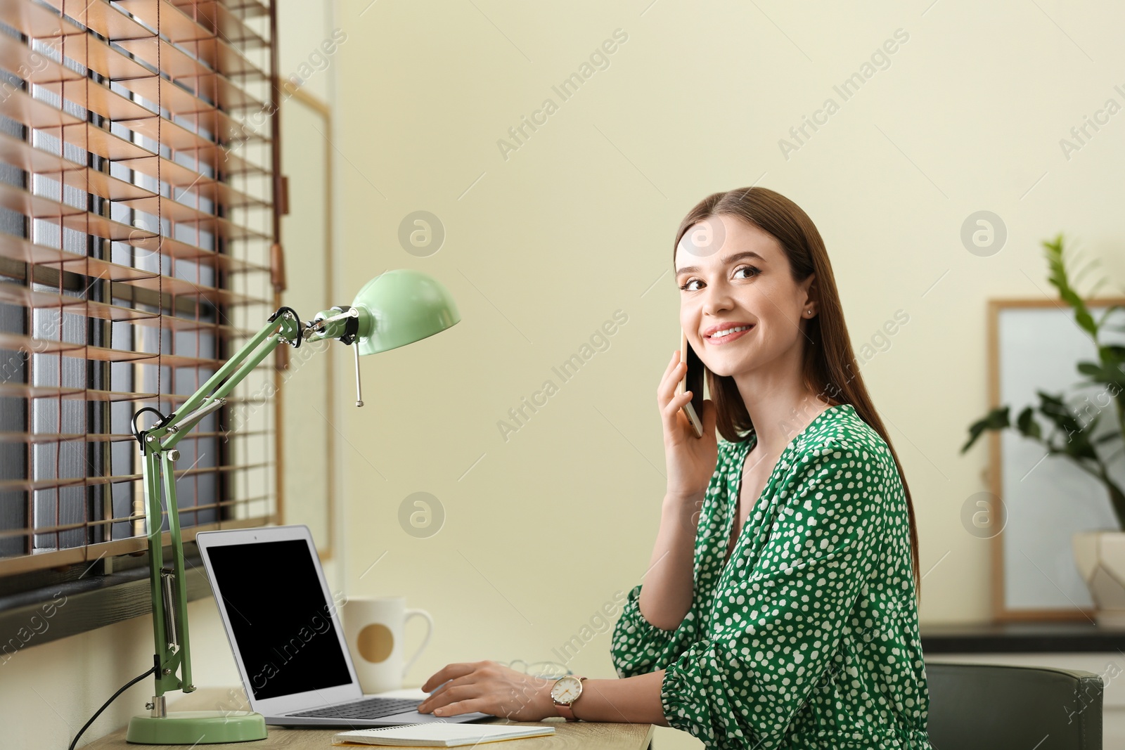 Photo of Young woman talking on phone while using laptop at table indoors. Space for text