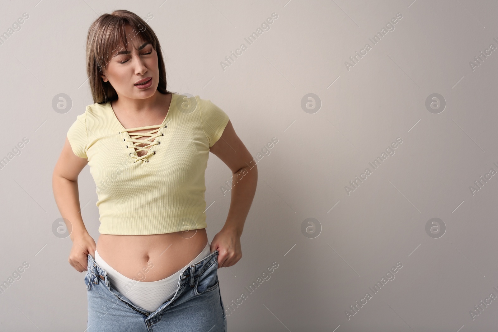 Photo of Young woman struggling to put on tight jeans against light grey background. Space for text