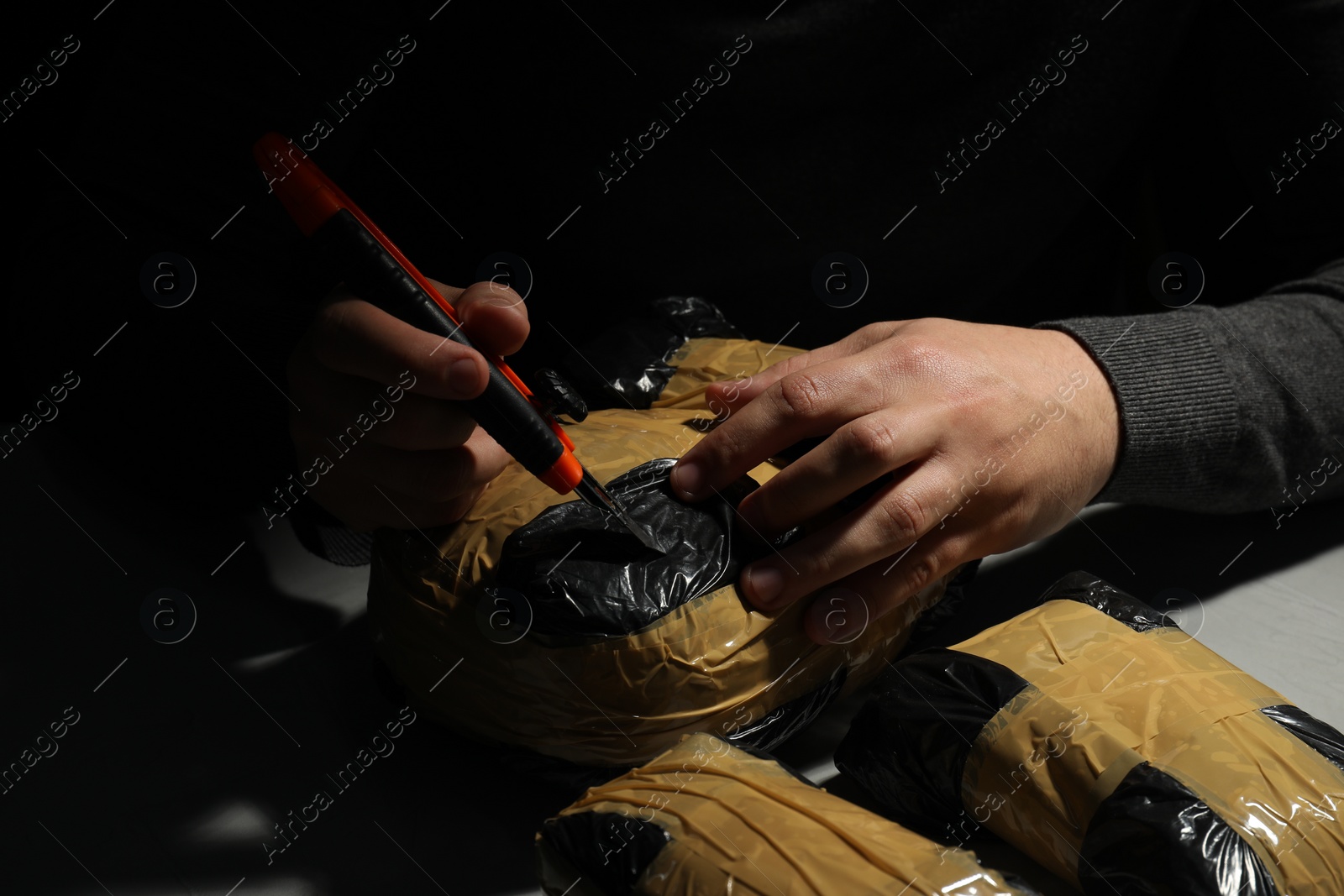 Photo of Smuggling and drug trafficking. Man opening package of narcotics with box cutter in darkness, closeup