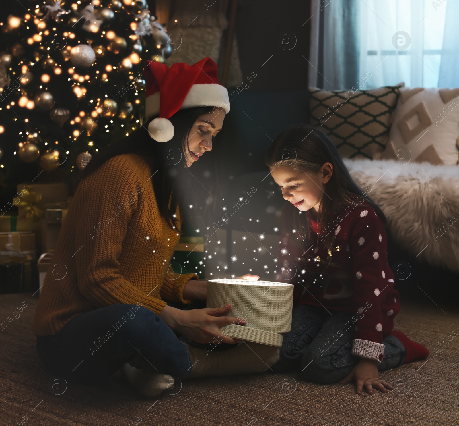 Image of Mother with her cute daughter opening gift box with magical light at home. Christmas celebration