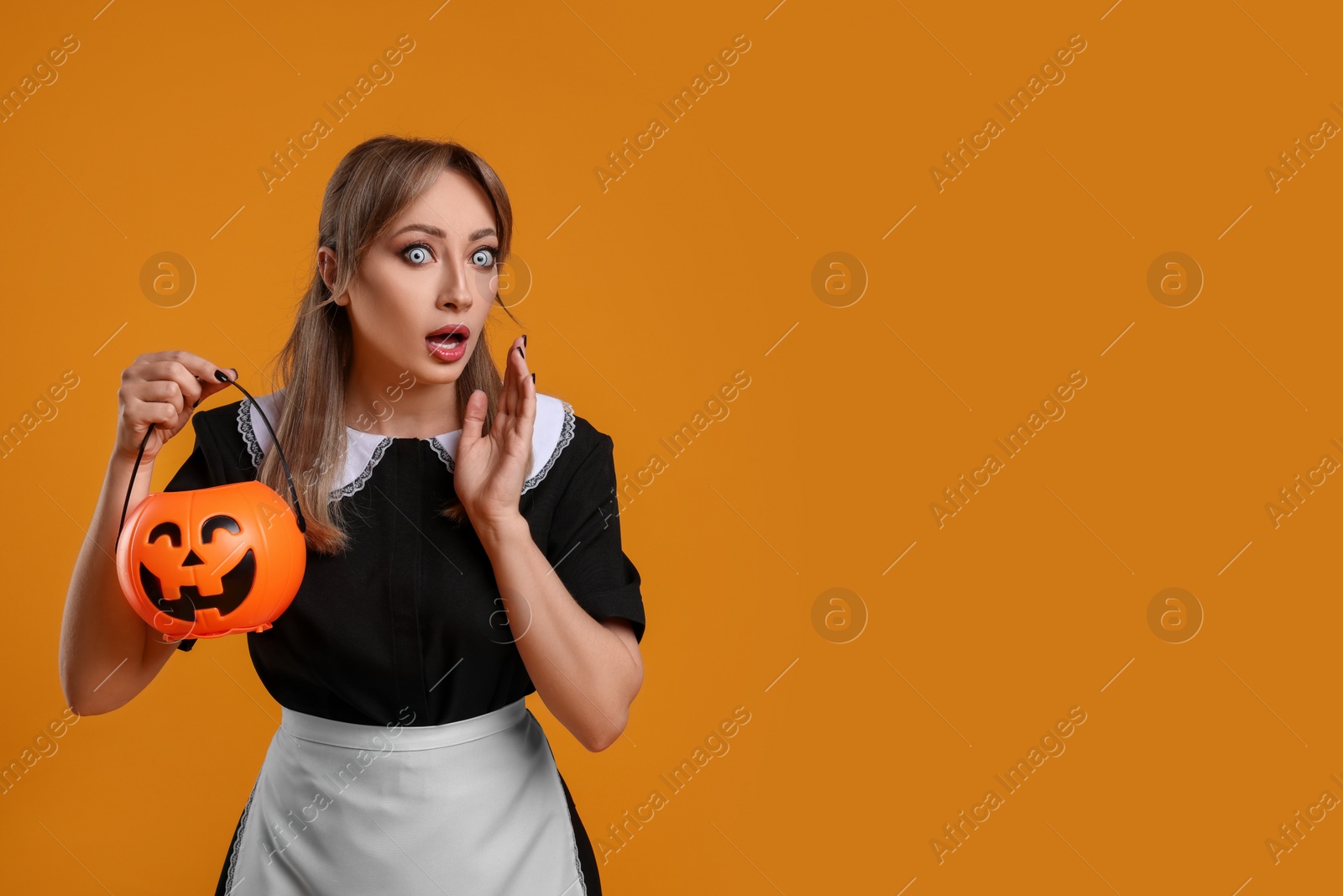 Photo of Emotional woman in scary maid costume with pumpkin bucket on orange background, space for text. Halloween celebration