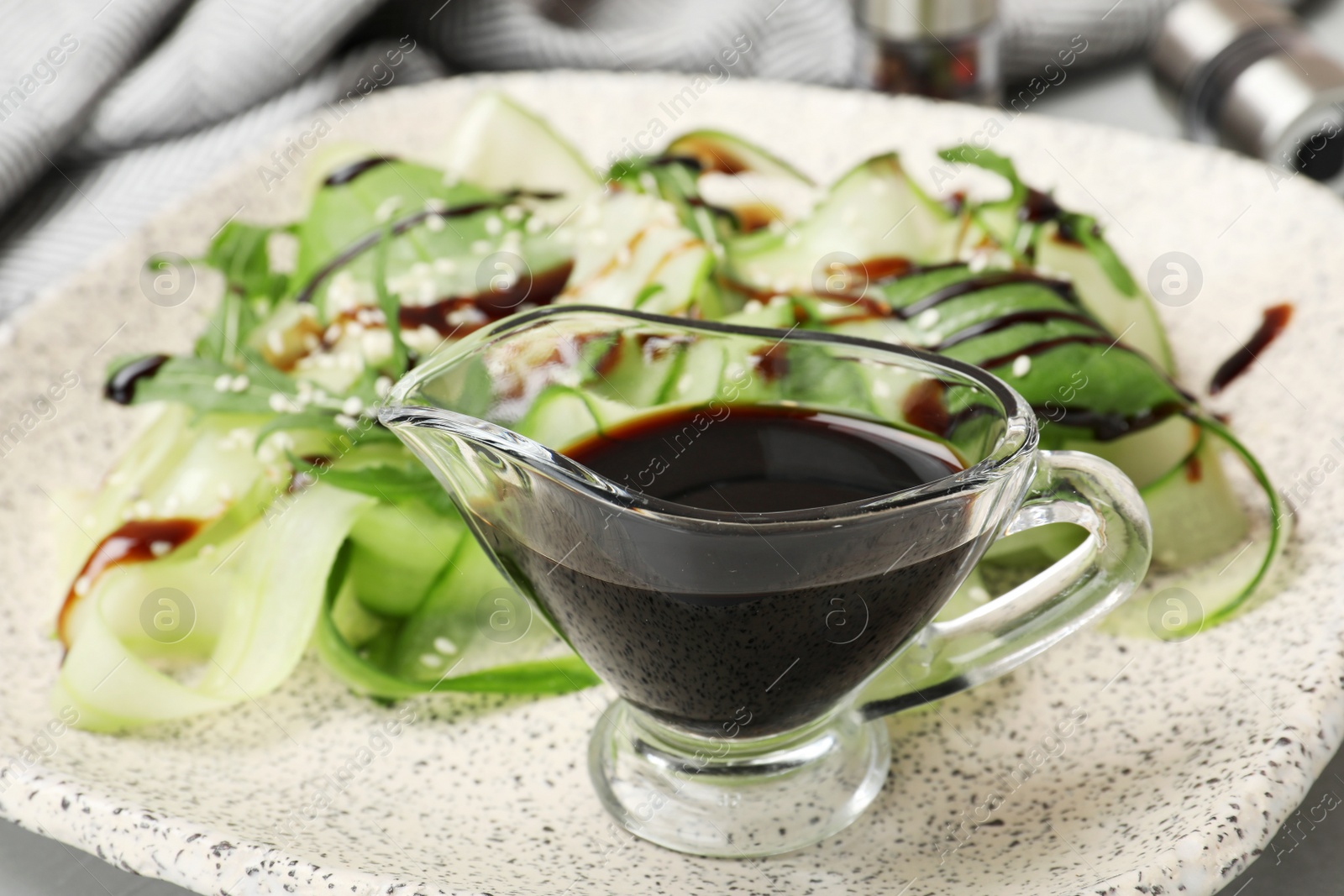 Photo of Gravy boat of balsamic vinegar on plate with vegetable salad, closeup