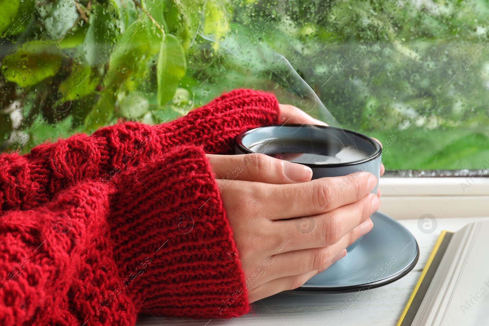 Photo of Woman with cup of hot drink near window on rainy day, closeup