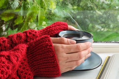 Photo of Woman with cup of hot drink near window on rainy day, closeup