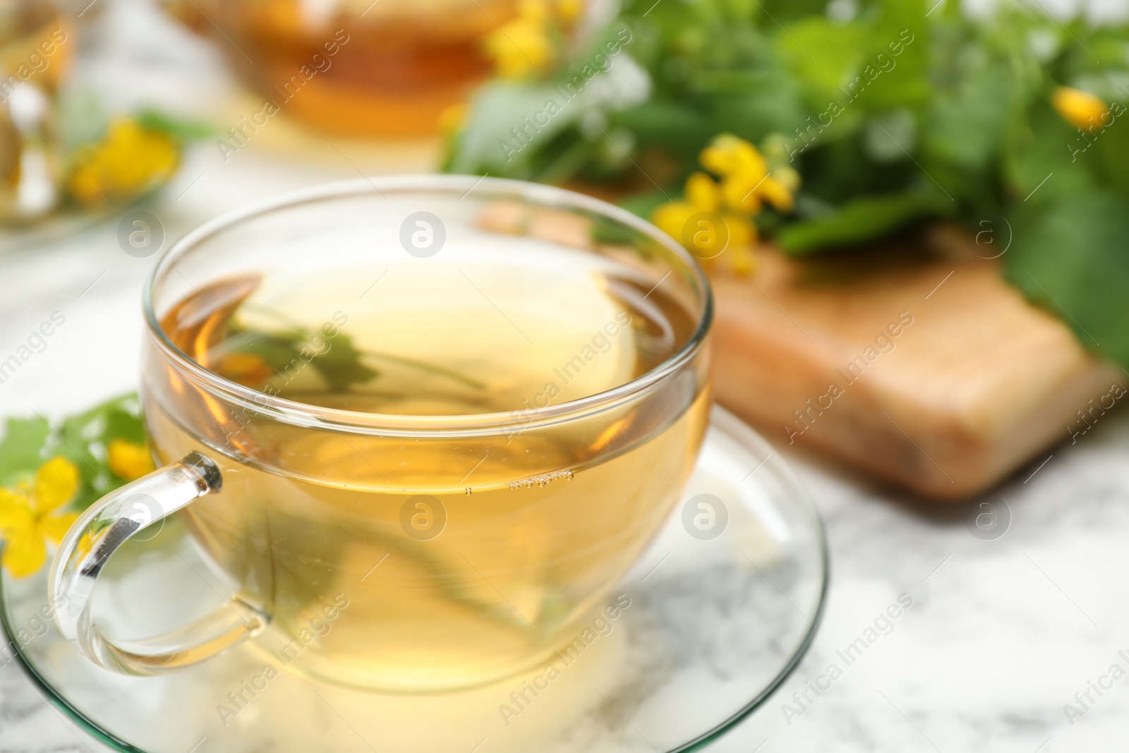 Photo of Glass cup of aromatic celandine tea and flowers on white table, closeup