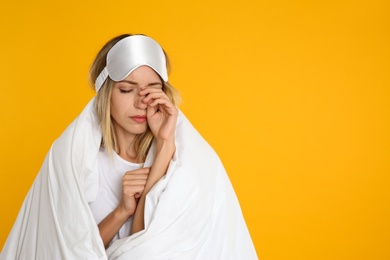 Young woman in sleeping mask wrapped with soft blanket on yellow background. Space for text