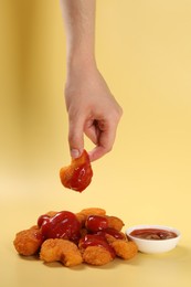 Woman holding delicious chicken nugget with ketchup on pale yellow background, closeup