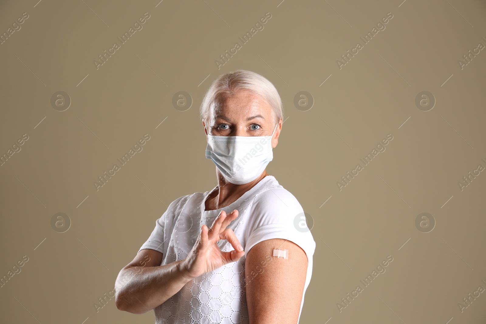 Photo of Mature woman in protective mask showing arm with bandage after vaccination and okay gesture on beige background