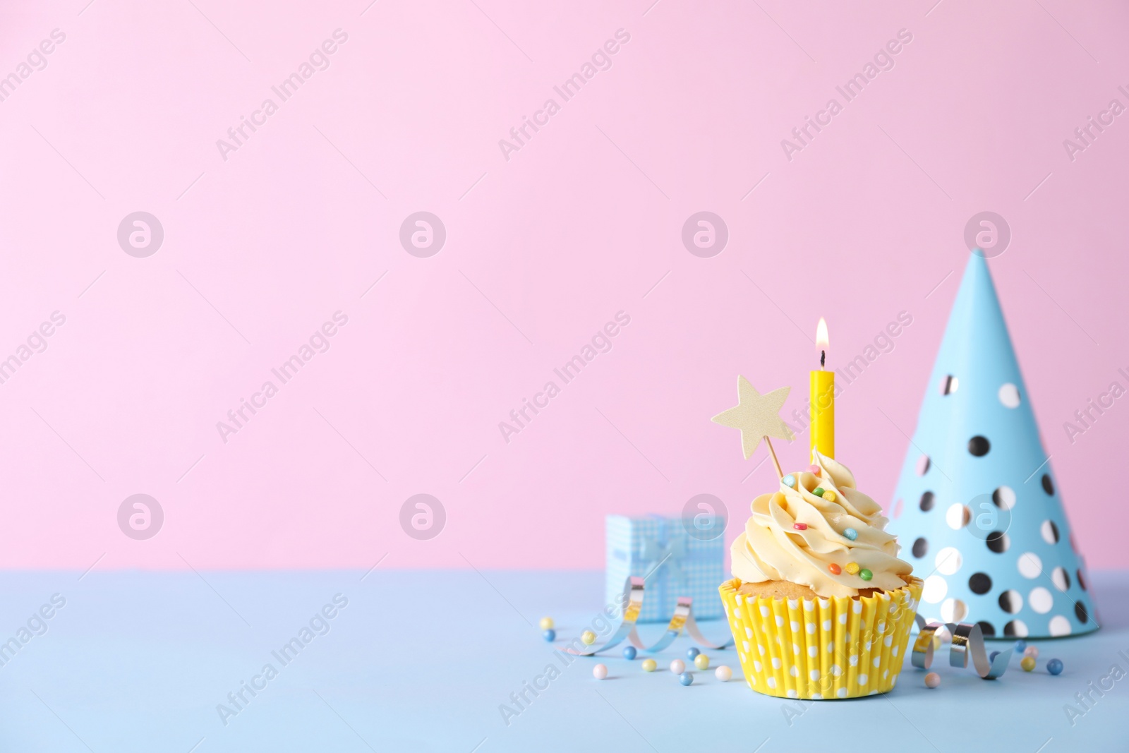 Photo of Delicious birthday cupcake with burning candle, party decor and gift box on light blue table against pink background, space for text