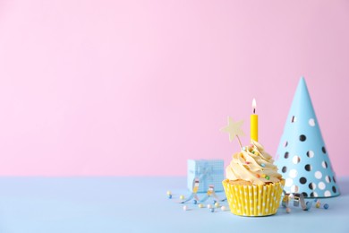 Photo of Delicious birthday cupcake with burning candle, party decor and gift box on light blue table against pink background, space for text