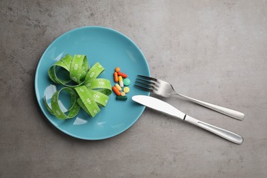 Plate with weight loss pills, measuring tape and cutlery on grey table, flat lay