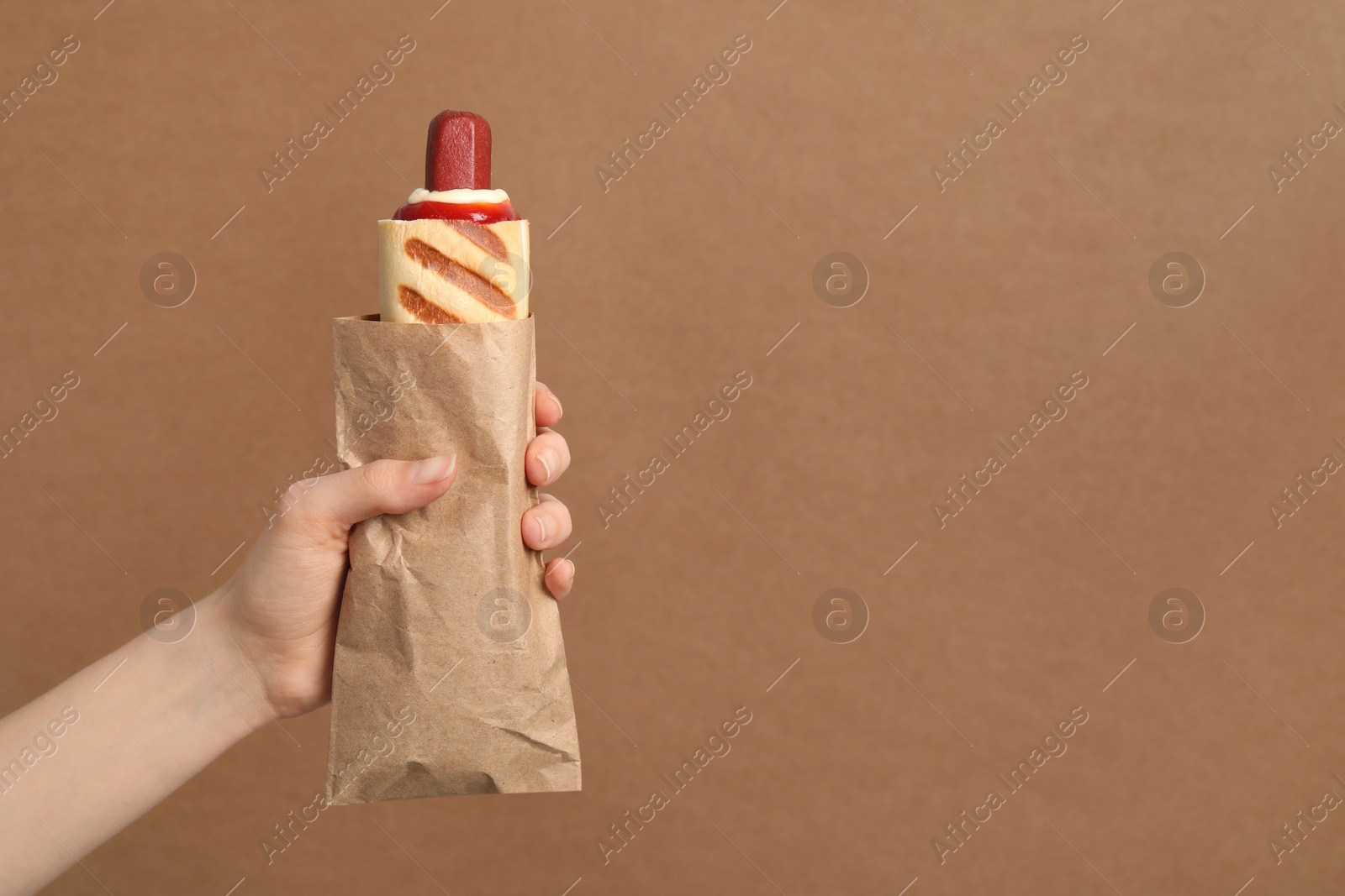 Photo of Woman holding paper bag with delicious french hot dog on brown background, closeup. Space for text