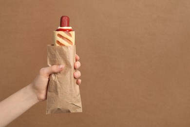 Photo of Woman holding paper bag with delicious french hot dog on brown background, closeup. Space for text