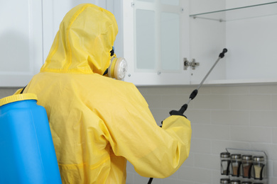 Pest control worker in protective suit spraying insecticide on furniture indoors