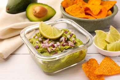 Photo of Bowl of delicious guacamole with onion, lime and nachos chips on white wooden table, closeup