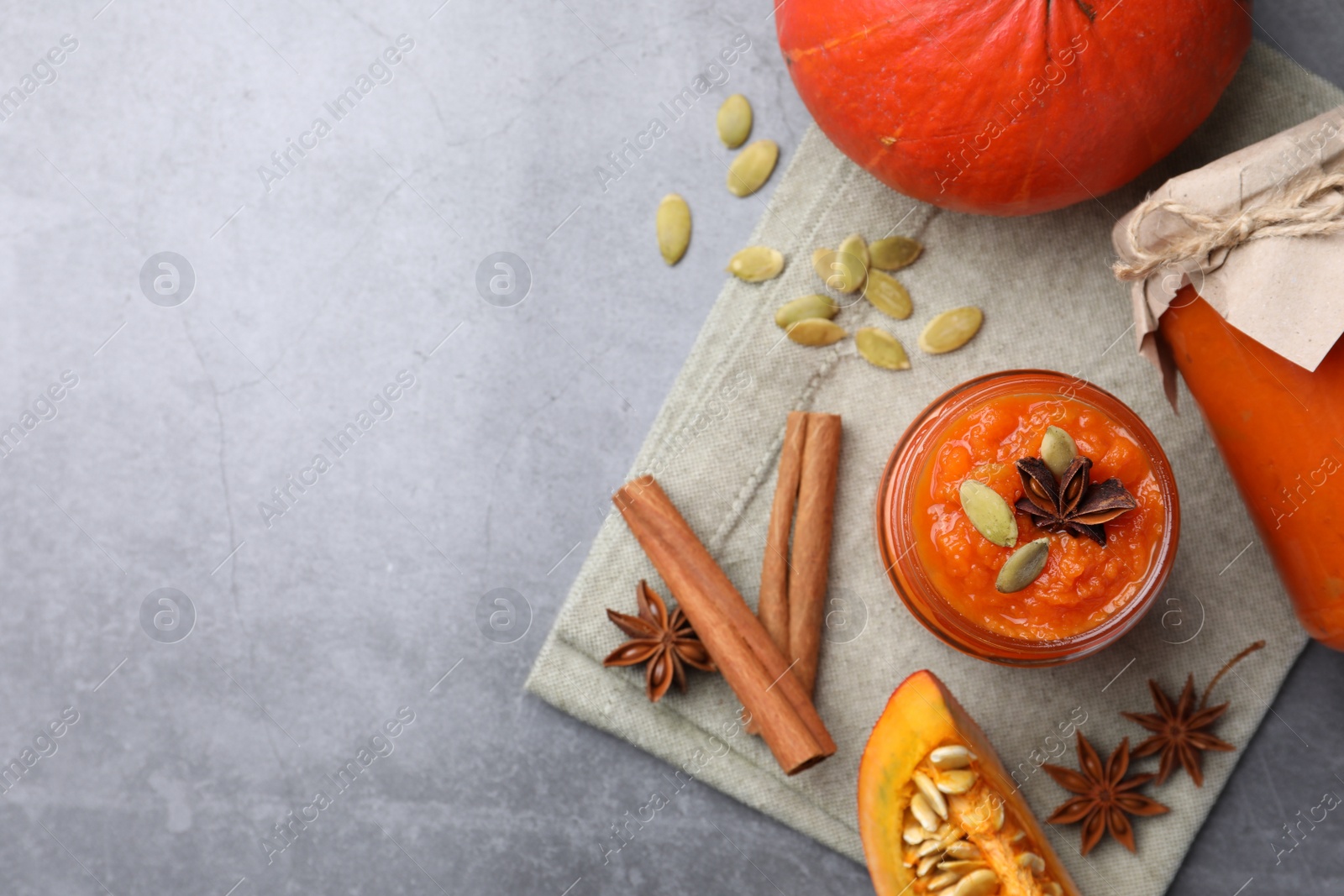 Photo of Jars of pumpkin jam, star anise, cinnamon and fresh pumpkins on grey table, flat lay. Space for text