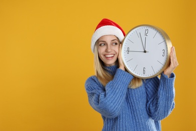 Photo of Woman in Santa hat with clock on yellow background, space for text. New Year countdown