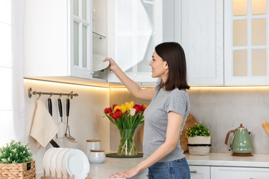 Photo of Beautiful young woman taking plate from kitchen cabinet at home