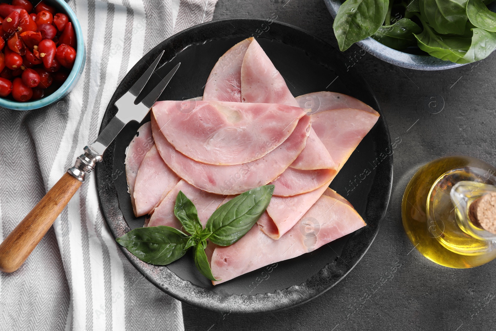 Photo of Tasty ham with basil, olive oil, pickled peppers and carving fork on grey table, flat lay