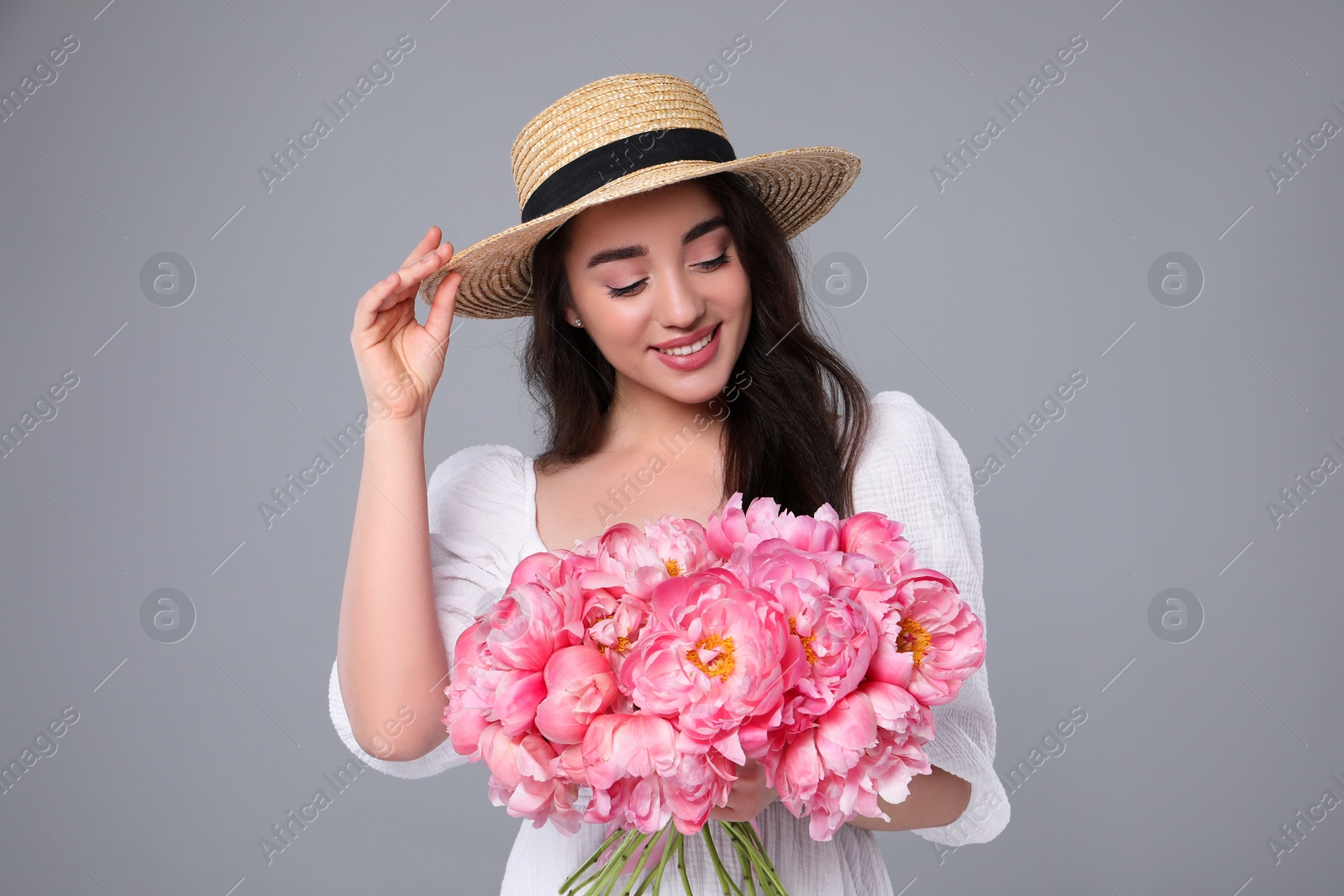 Photo of Beautiful young woman in straw hat with bouquet of pink peonies against grey background