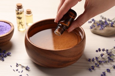 Photo of Woman dripping lavender essential oil from bottle into bowl at white wooden table, closeup