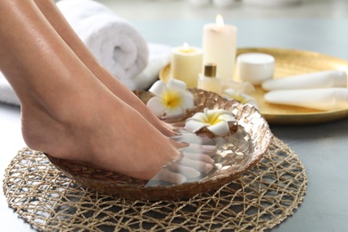 Closeup view of woman soaking her feet in dish with water and flowers on grey floor. Spa treatment