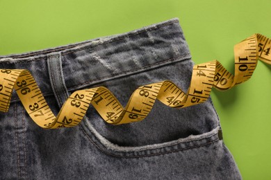 Jeans with measuring tape on green background, top view. Weight loss concept