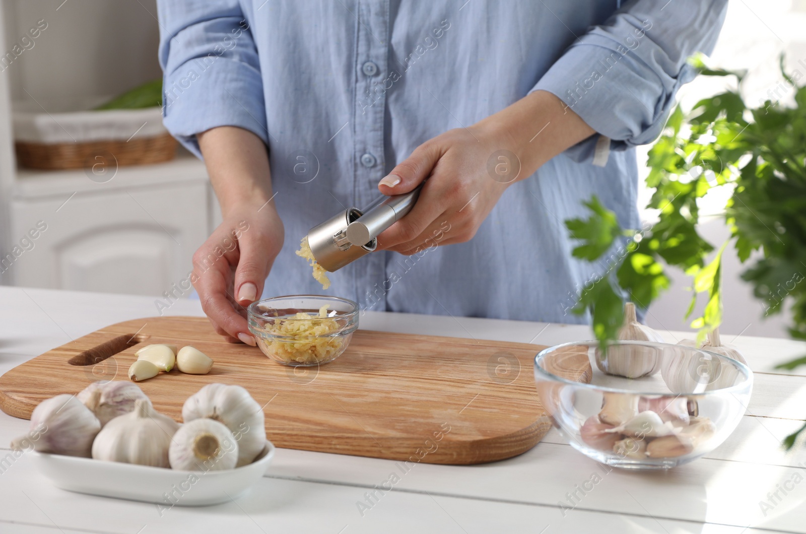 Photo of Woman squeezing garlic with press at white wooden table in kitchen, closeup