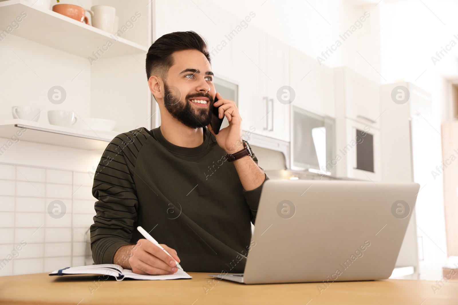 Photo of Handsome young man talking on phone while working at table in kitchen