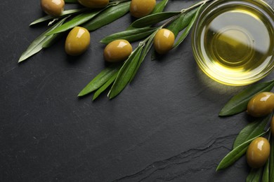 Photo of Cooking oil in bowl, olives and leaves on black textured table, flat lay. Space for text