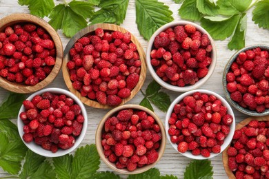 Photo of Fresh wild strawberries in bowls and leaves on white wooden table, flat lay