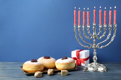 Photo of Silver menorah, dreidels with He, Pe, Nun, Gimel letters, gift box and sufganiyot on blue wooden table, space for text. Hanukkah symbols