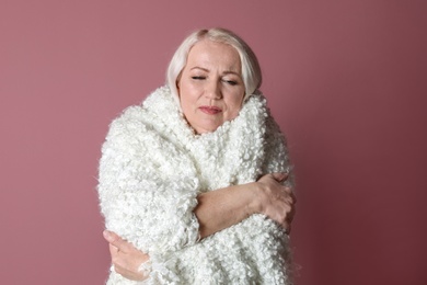 Mature woman wrapped in blanket suffering from cold on color background