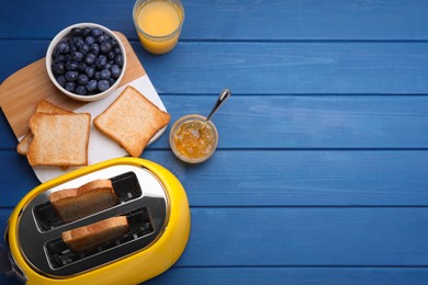 Photo of Yellow toaster with roasted bread, glass of juice, blueberries and jam on blue wooden table, flat lay. Space for text