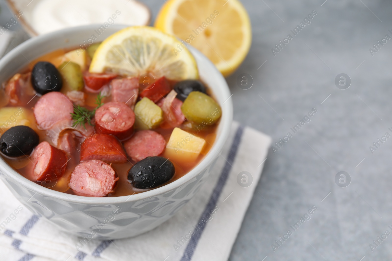 Photo of Meat solyanka soup with thin dry smoked sausages in bowl on grey table, closeup. Space for text