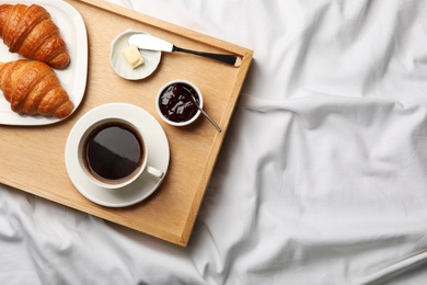 Top view of wooden tray with coffee, croissants, butter and jam on white fabric, space for text. Breakfast in bed