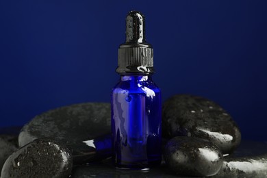 Photo of Bottle of face serum with wet spa stones on blue background, closeup