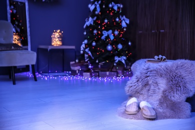 Photo of Soft slippers with fluffy blanket and Christmas tree in dark room