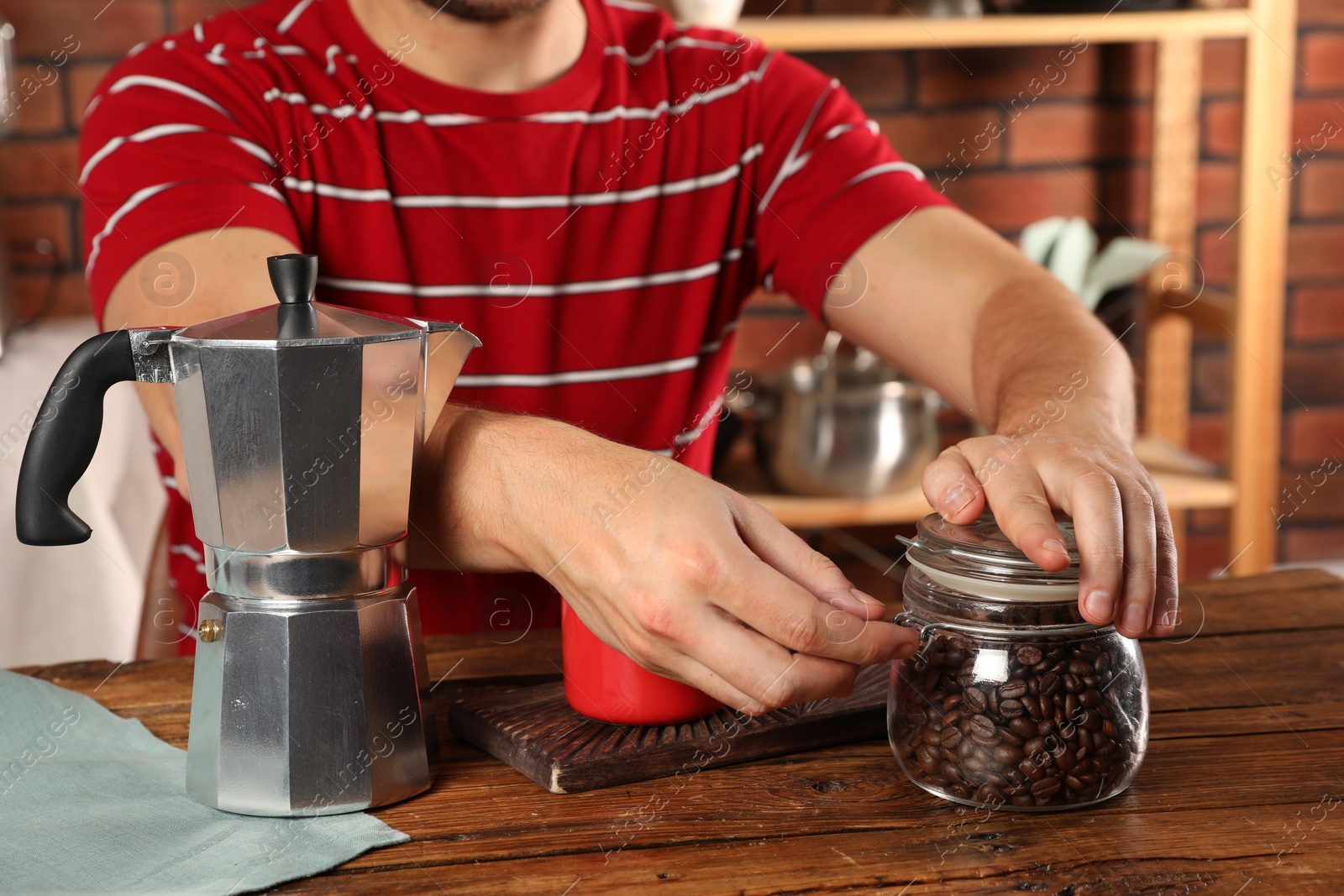 Photo of Brewing coffee. Man with jar of beans, moka pot and mug at wooden table indoors, closeup