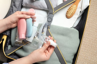 Photo of Woman putting small bottle with personal care product into plastic bag near suitcase, closeup. Cosmetic travel kit