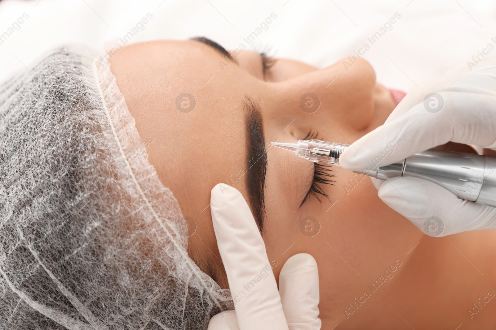 Photo of Young woman undergoing procedure of permanent eyebrow makeup in tattoo salon, closeup